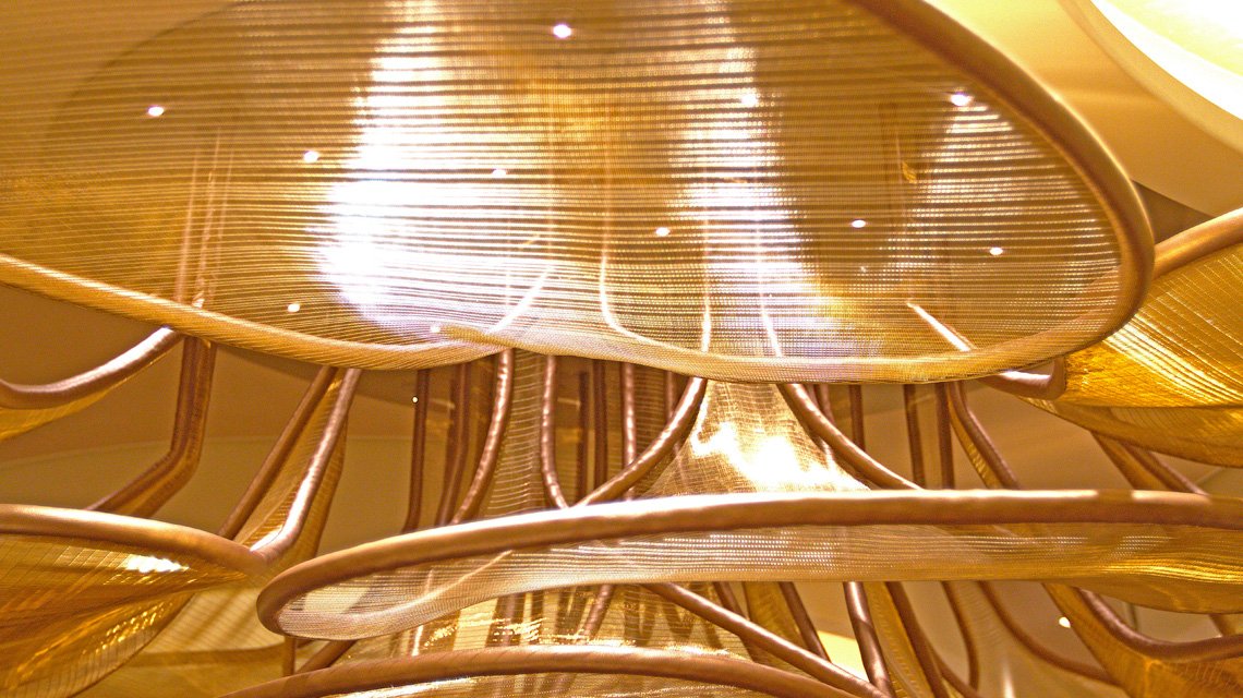 Detalhe da malha metálica Liner Gold - <p>Vista de detalhe da malha metálica arquitetónica Liner Gold utilizada na criação do lustre monumental do restaurante Le Voyage. A textura e os reflexos dourados conferem elegância e leveza ao projeto.</p>